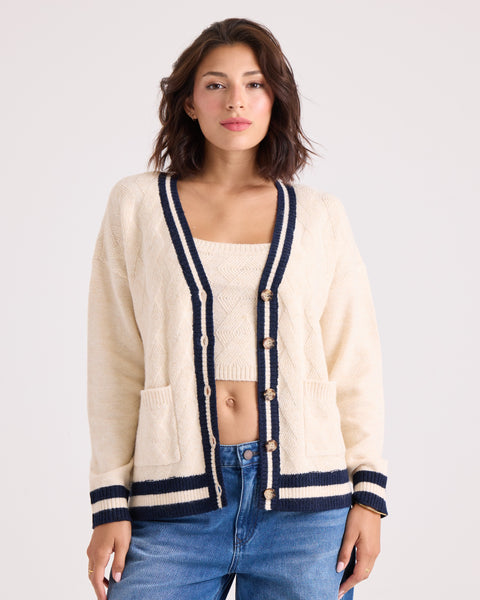 A woman is wearing the Volcom Women's Boyfriend Cardigan in cream with black trim and dropped shoulders, layered over a matching top and paired with blue jeans. She stands against a plain light background, looking at the camera with a neutral expression.