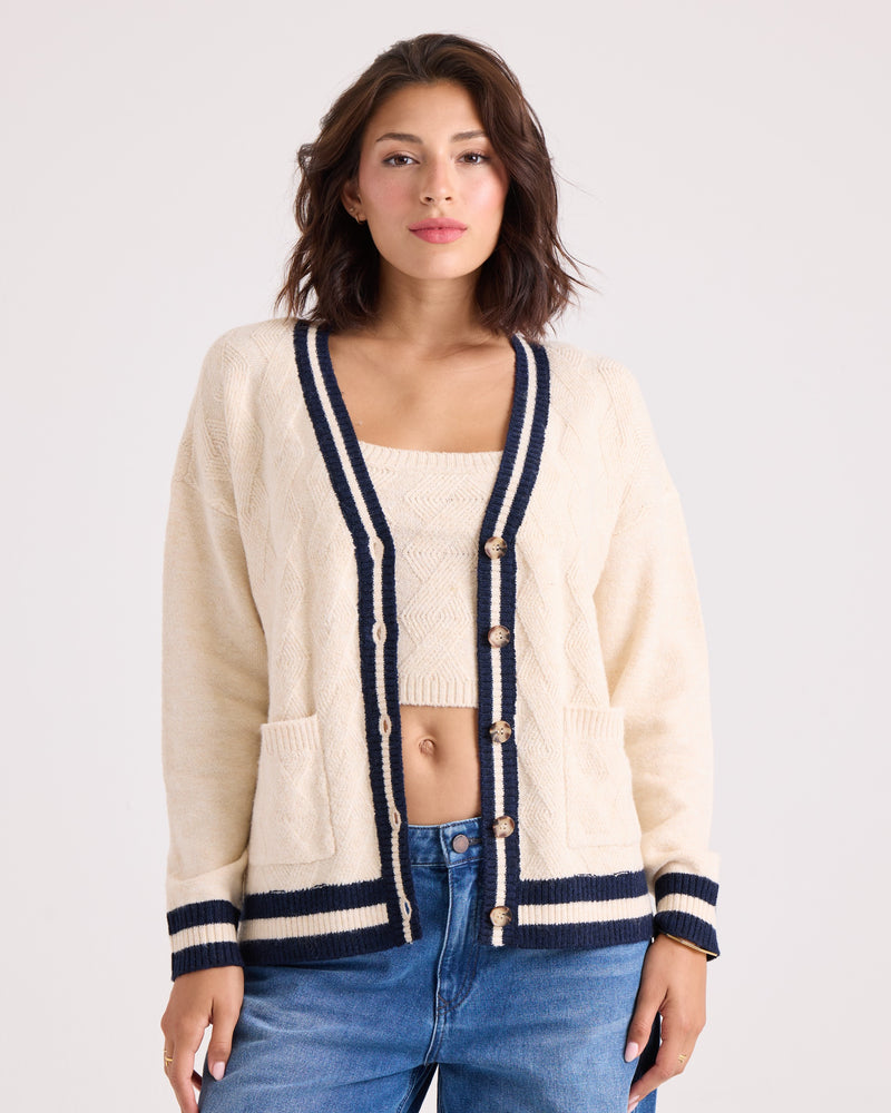 A woman is wearing the Volcom Women's Boyfriend Cardigan in cream with black trim and dropped shoulders, layered over a matching top and paired with blue jeans. She stands against a plain light background, looking at the camera with a neutral expression.