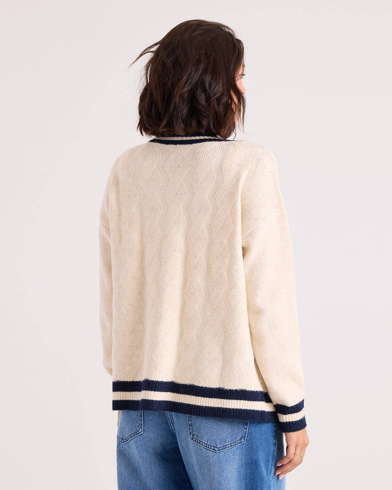 A woman with short brown hair, dressed in the Volcom Women's Boyfriend Cardigan in cream with black striped cuffs and collar, dropped shoulders, and blue jeans, stands facing away from the camera against a light background.