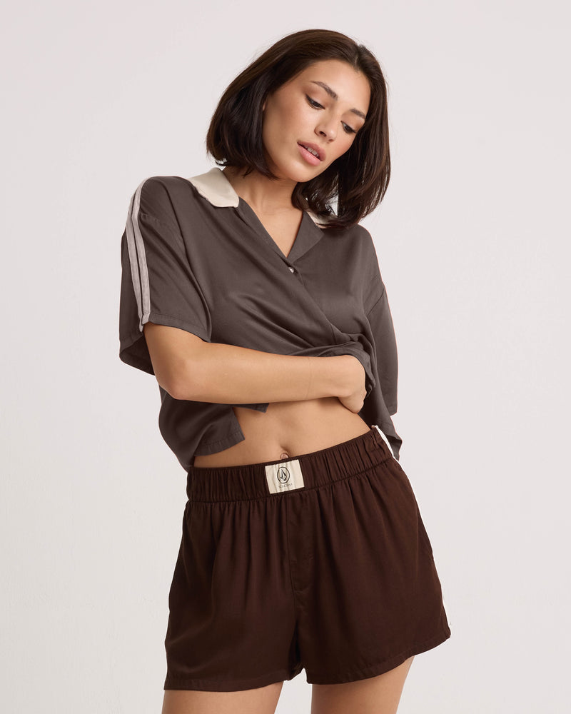 A woman with shoulder-length brown hair wears the Volcom Women's Boxing Shirt, featuring a loose, cropped silhouette, paired with matching brown shorts. She stands against a plain white background, lifting her shirt slightly with one hand.