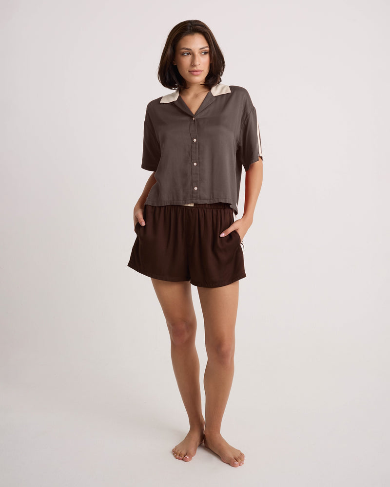 A woman with short dark hair is barefoot, standing against a plain white background with her hands in her pockets, wearing the Volcom Women's Boxing Shirt—featuring a cropped brown button-up design with a light collar—and matching brown shorts, looking slightly to the side.