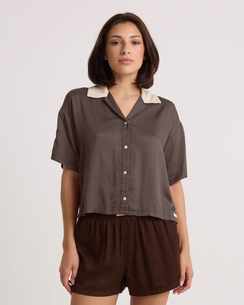 A woman with shoulder-length brown hair is wearing the Volcom Women's Boxing Shirt— a cropped, short-sleeved, button-up brown top with a white collar—paired with matching brown shorts, standing against a plain white background.