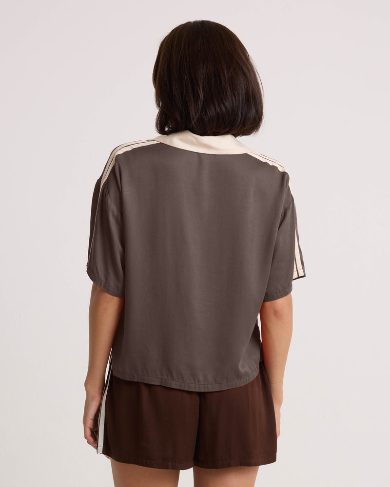 A woman with shoulder-length dark hair wears the Volcom Women's Boxing Shirt, a loose brown top with cream trim and a cropped silhouette, paired with matching brown shorts. She stands with her back to the camera against a plain, light background.