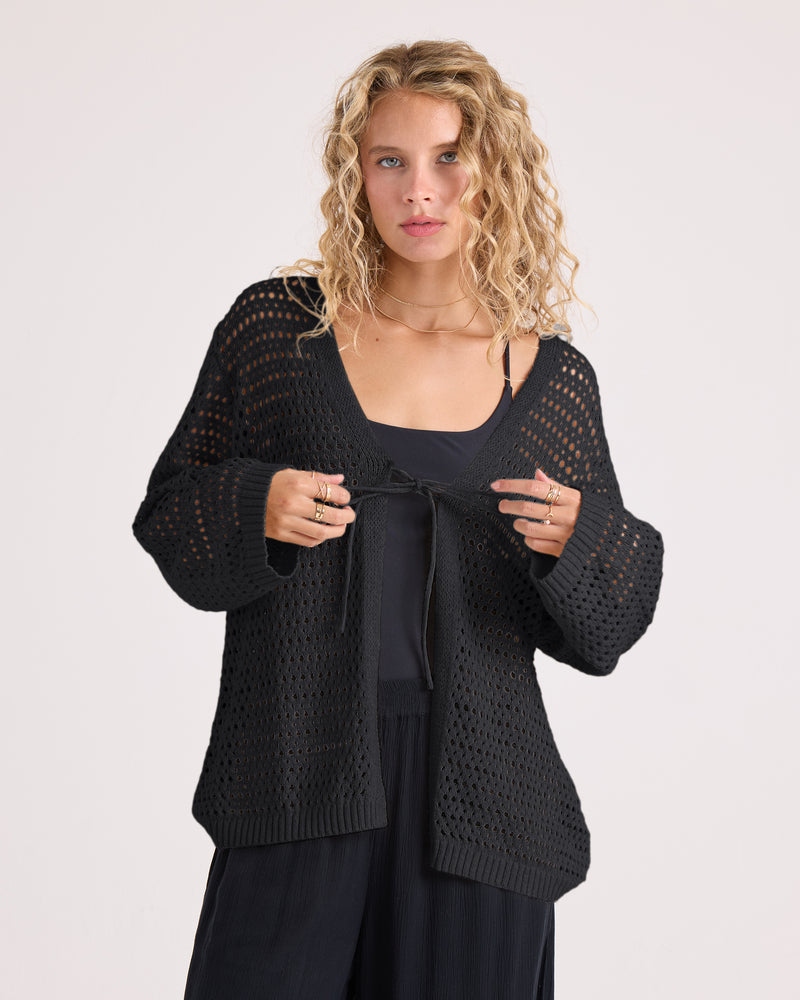A woman with curly blonde hair wears the Volcom Women’s Go Go Wrap, a black soft knit cardigan, over a black top and black pants as she stands against a plain light background.