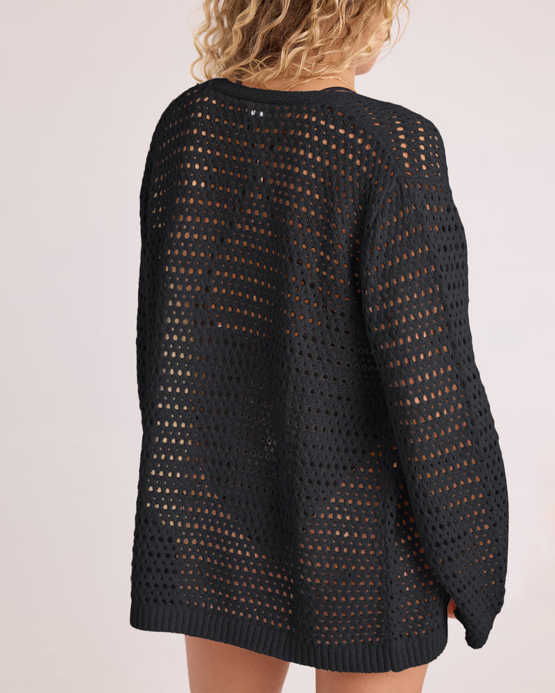 A person with curly hair is seen from the back wearing the Volcom Women’s Go Go Wrap, a loose, black, open-knit tie front cardigan. The sheer garment reveals a black undergarment and bare legs against a plain, light-colored background.