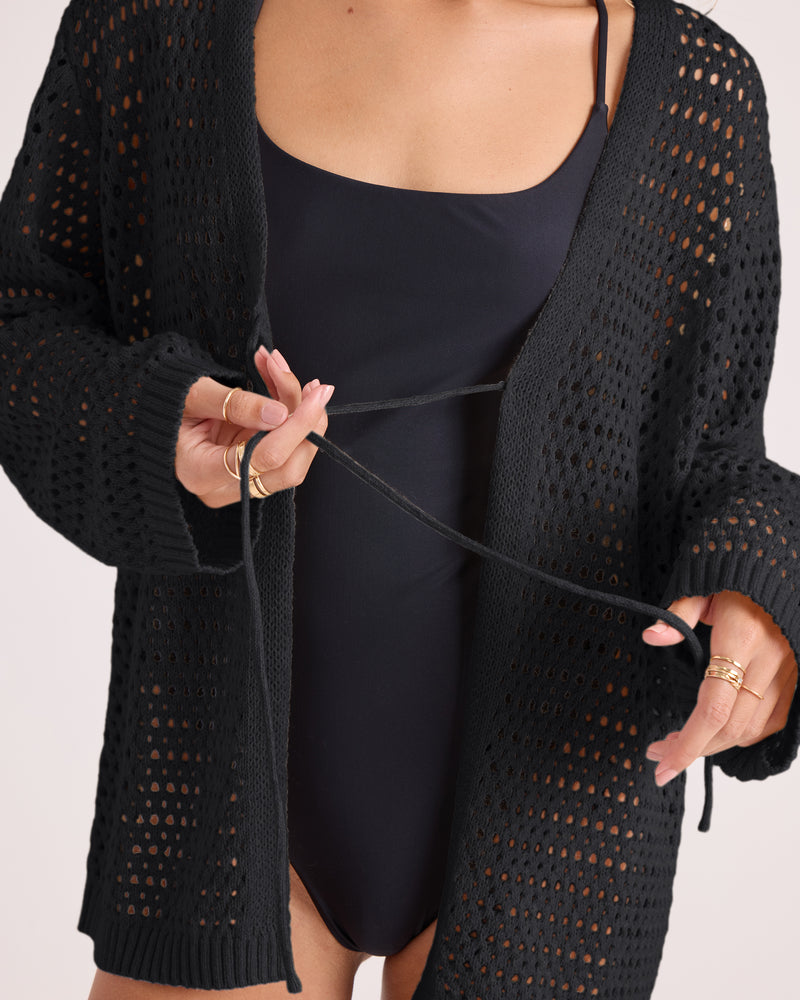 A person wearing a black swimsuit styles the Volcom Women’s Go Go Wrap, a soft knit tie front cardigan, by tying it at the waist. They have light pink nails and wear several gold rings. The background is plain and light-colored.