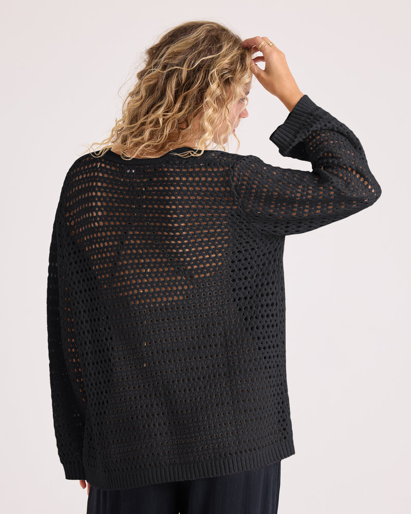 A person with wavy blond hair is seen from behind, wearing the Volcom Women’s Go Go Wrap—a soft, black open-knit sweater—and touching their head with one hand. The background is plain and light-colored.