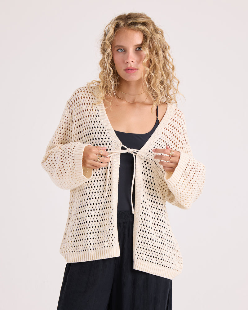 A woman with curly blonde hair wears the Volcom Women’s Go Go Wrap—a loose, cream-colored cotton-acrylic blend crochet cardigan—over a black top and black pants, standing against a plain light background.