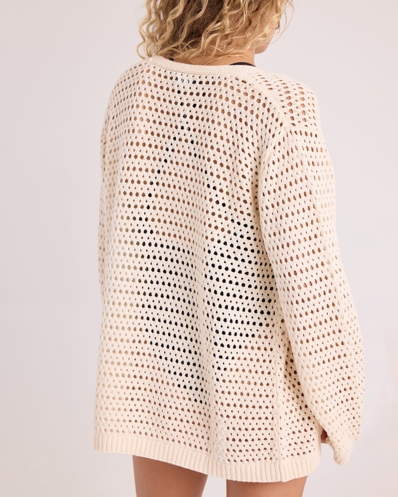 A person with curly blonde hair is photographed from behind against a plain light background, wearing the Volcom Women’s Go Go Wrap—a loose, cream-colored, open-knit sweater top.