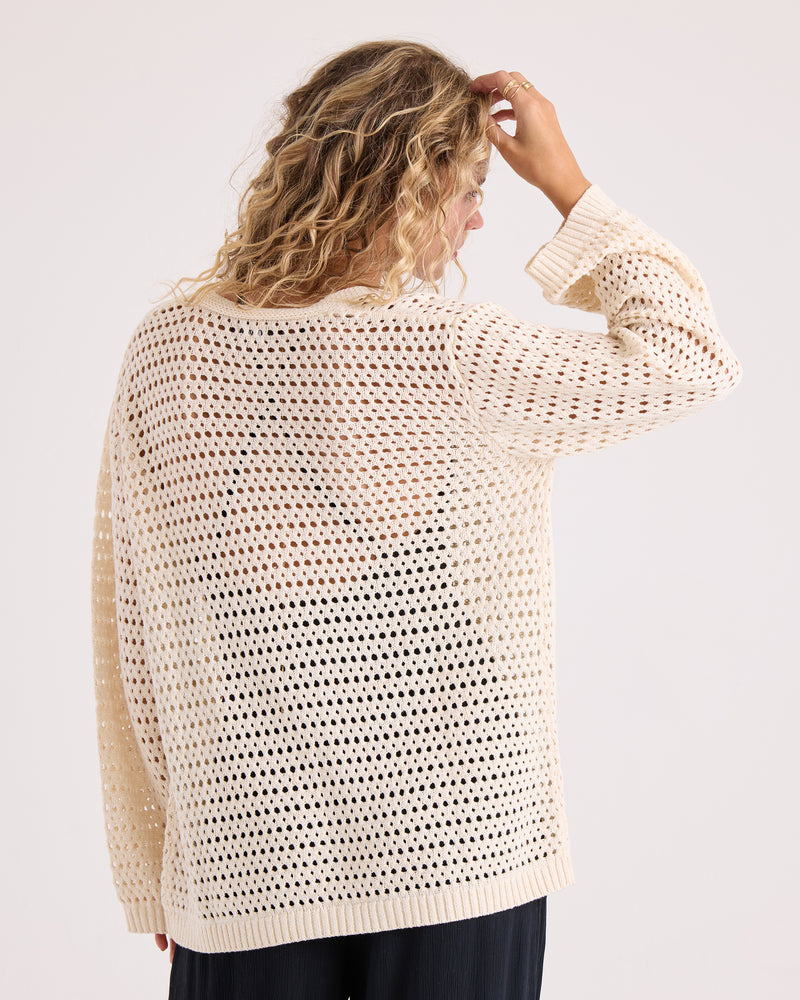 A person with wavy blonde hair is wearing the Volcom Women’s Go Go Wrap—a loose, cream-colored, open-knit sweater top crafted from a soft cotton-acrylic blend—paired with black pants. They are facing away from the camera and touching their head with one hand against a plain, light background.