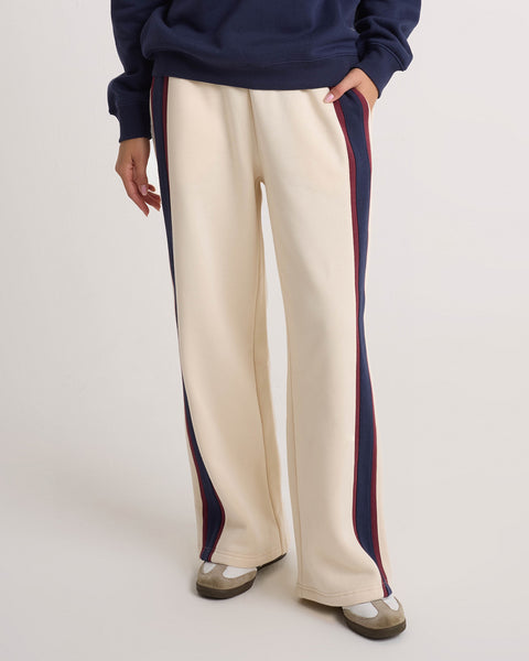 A person is wearing Volcom Women's Wide Leg Sweatpants in a cream color with navy and red side stripes, featuring an elastic waistband and relaxed fit, paired with a navy top and white sneakers with brown details, standing against a plain background.