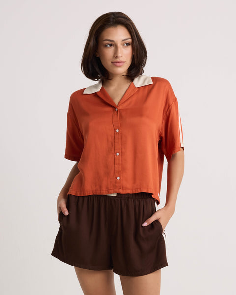 A woman with short brown hair stands against a plain background, looking to the side with one hand in her pocket. She wears a lightweight orange collared short-sleeve shirt paired with Volcom Women's Boxing Shorts—brown shorts featuring an elastic waistband.