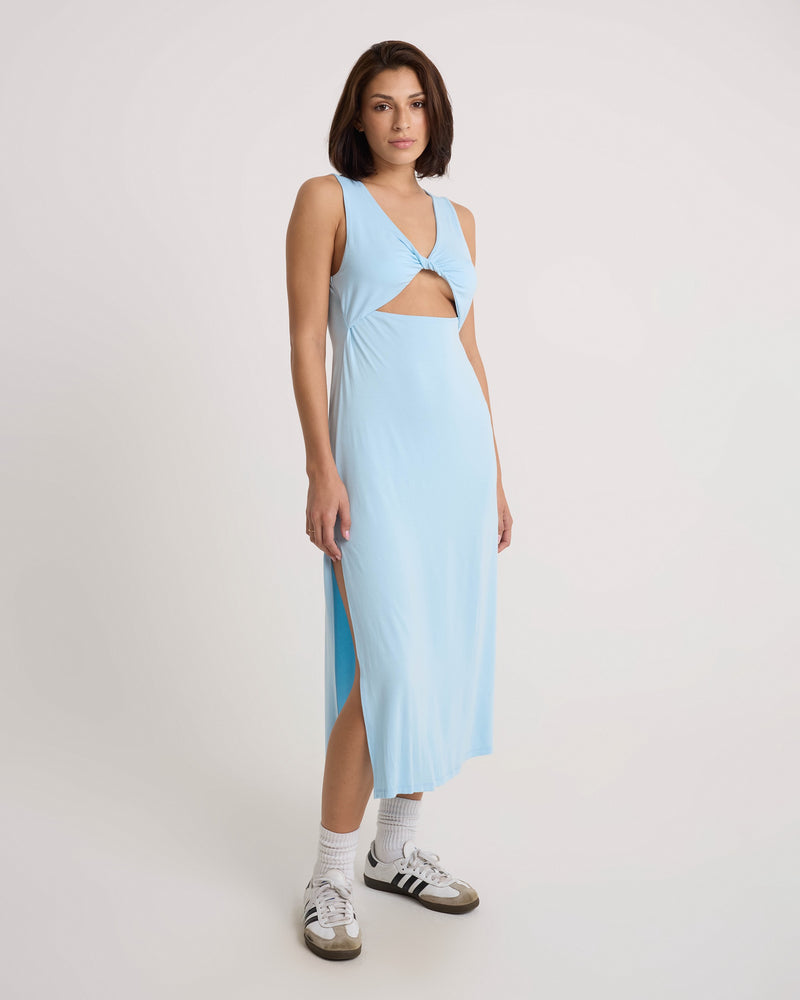 A woman stands against a plain backdrop wearing the Volcom Women's Stone Luz Dress, which features a sleeveless twist-front bodice and side slit. She pairs it with white socks and black Adidas sneakers.