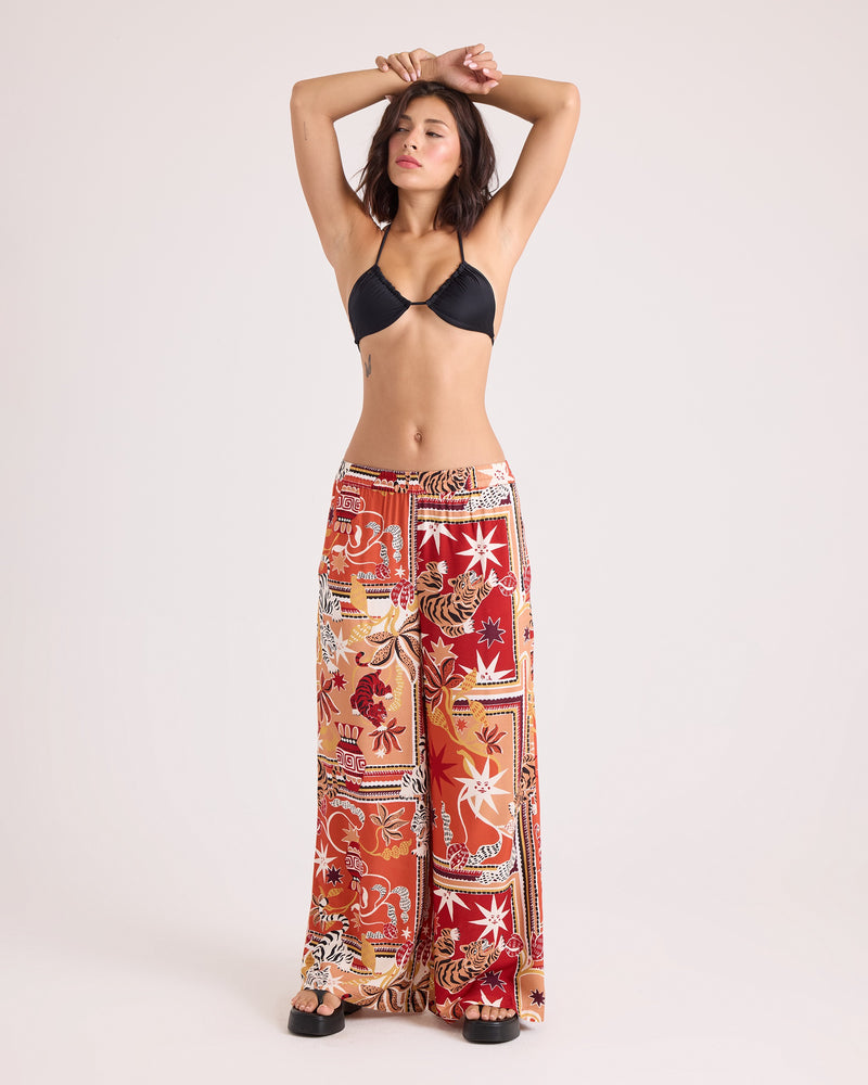 A woman stands with arms raised, wearing a black bikini top and Volcom Women's Stoneshine Junki Pants—relaxed fit, wide-leg pants featuring a red, orange, and white floral patchwork pattern with an elastic waistband—against a plain light background.