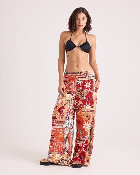 A woman stands against a light background wearing a black bikini top and the Volcom Women's Stoneshine Junki Pants, which feature a vibrant red and orange pattern, wide-leg silhouette, and elastic waistband. She has a neutral expression and relaxed posture.