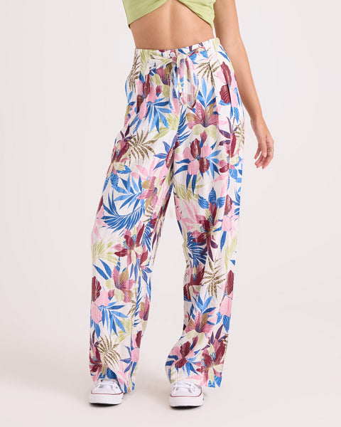 Wearing the Volcom Women's Bow Chicka Pants—loose, high-waisted, wide-leg trousers in a colorful tropical leaf and flower print with a relaxed fit and elastic waistband—paired with a light green crop top and white sneakers, the person stands against a plain background.