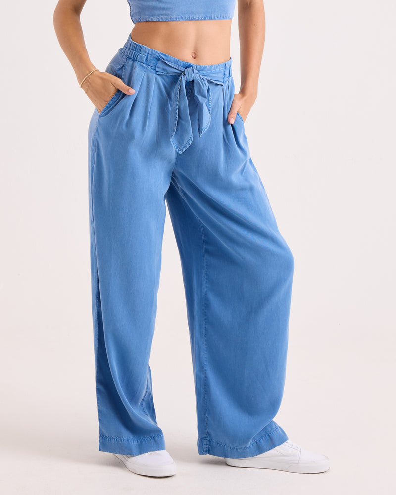 A person wearing Volcom’s Women’s Bow Chicka Pants—loose-fitting, high-waisted cerulean wide-leg woven trousers featuring an elastic waistband and matching belt—paired with a blue crop top and white sneakers, stands against a plain light background.