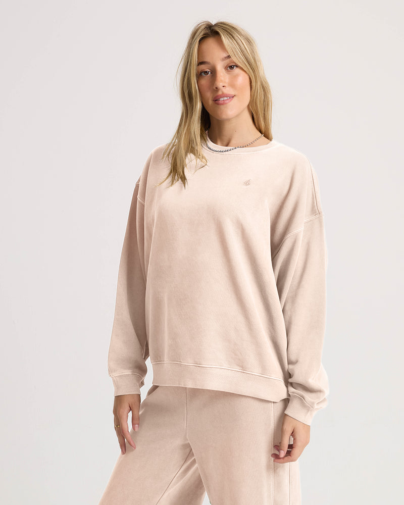 A woman stands against a plain background, looking at the camera with a relaxed expression while wearing the Volcom Women’s Stone Soft Crew in light beige slub brushed back fleece and matching sweatpants from the Lived In Lounge collection.