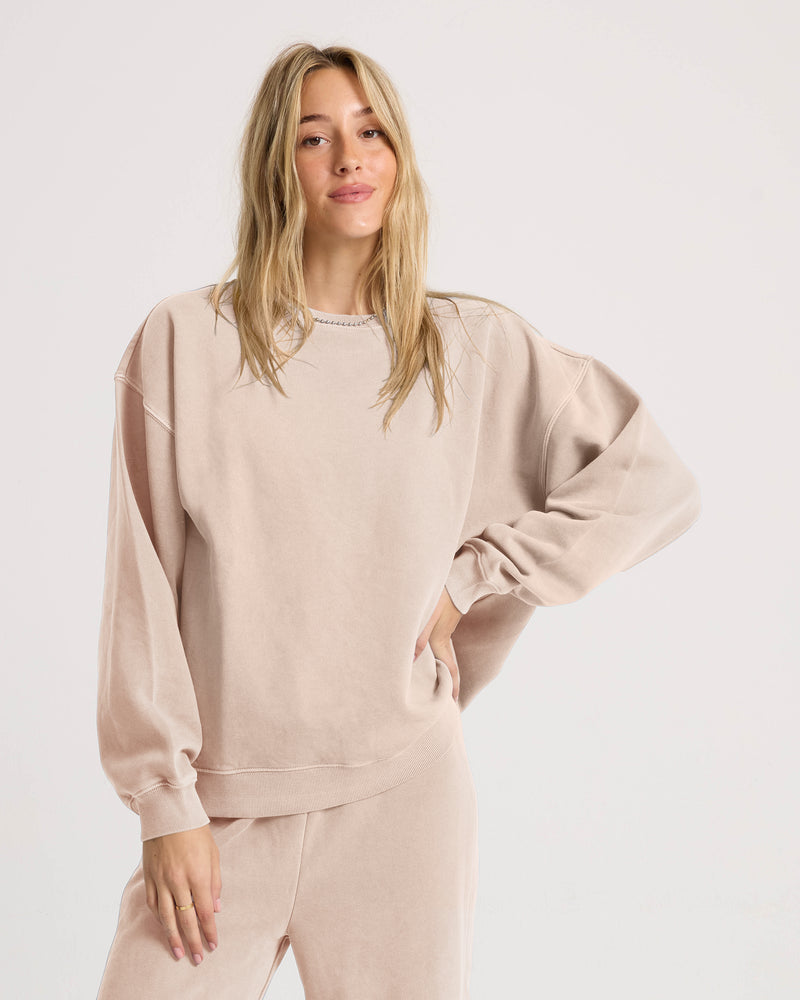 A woman with long blonde hair wears a loose, light beige Volcom Women’s Stone Soft Crew crafted from slub brushed back fleece, paired with matching pants. She stands with one hand on her hip against a plain white background—perfectly embodying the effortless comfort of Volcom’s Lived In Lounge collection.