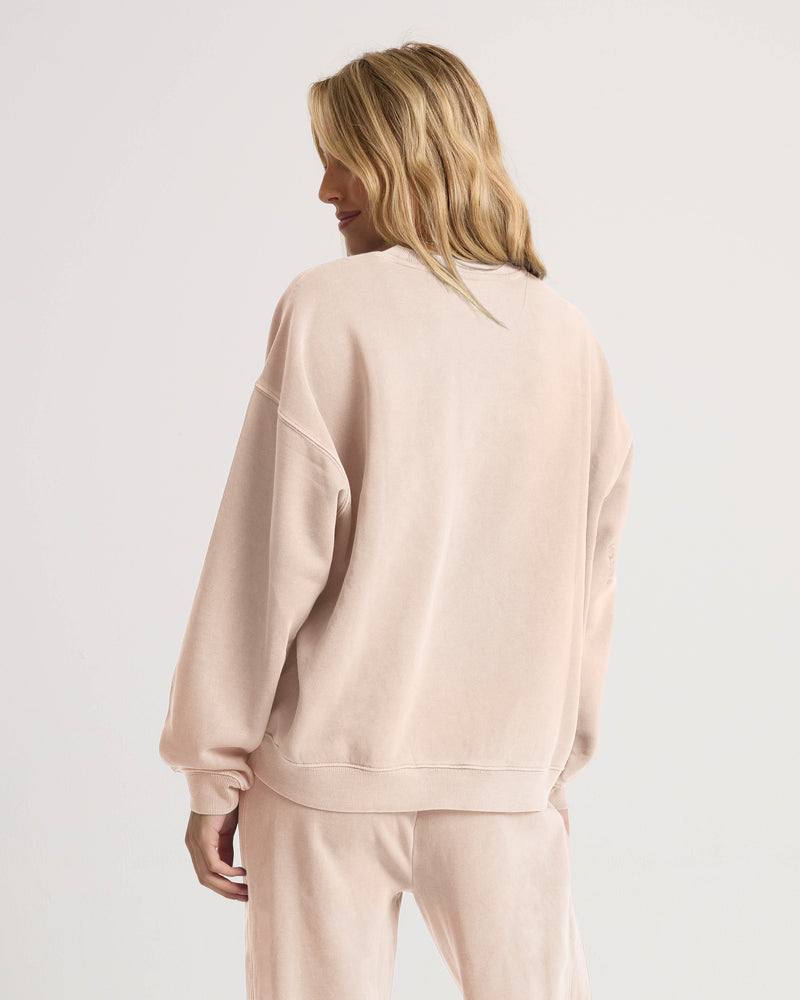 A woman with wavy blonde hair stands turned away, wearing the Volcom Women’s Stone Soft Crew in light beige—crafted from slub brushed back fleece—paired with matching pants against a plain white background, embodying the perfect look for the Lived In Lounge.