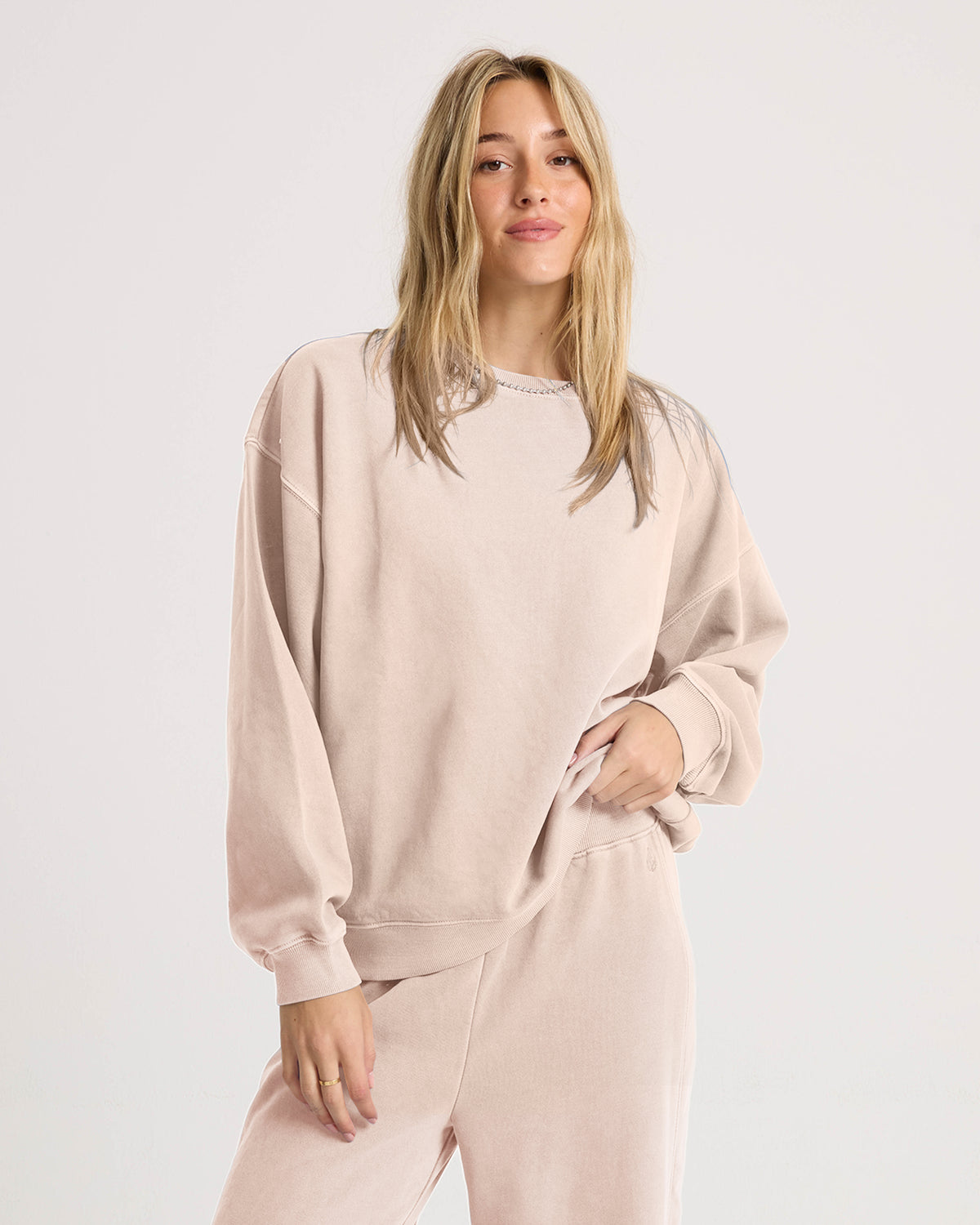 A woman with long blonde hair stands against a plain light background, softly smiling with one hand in her pocket as she wears the Volcom Women’s Stone Soft Crew sweatshirt and matching pants in light beige. Crafted from ultra-cozy slub brushed back fleece, her effortlessly relaxed look captures the signature Lived In Lounge vibe.