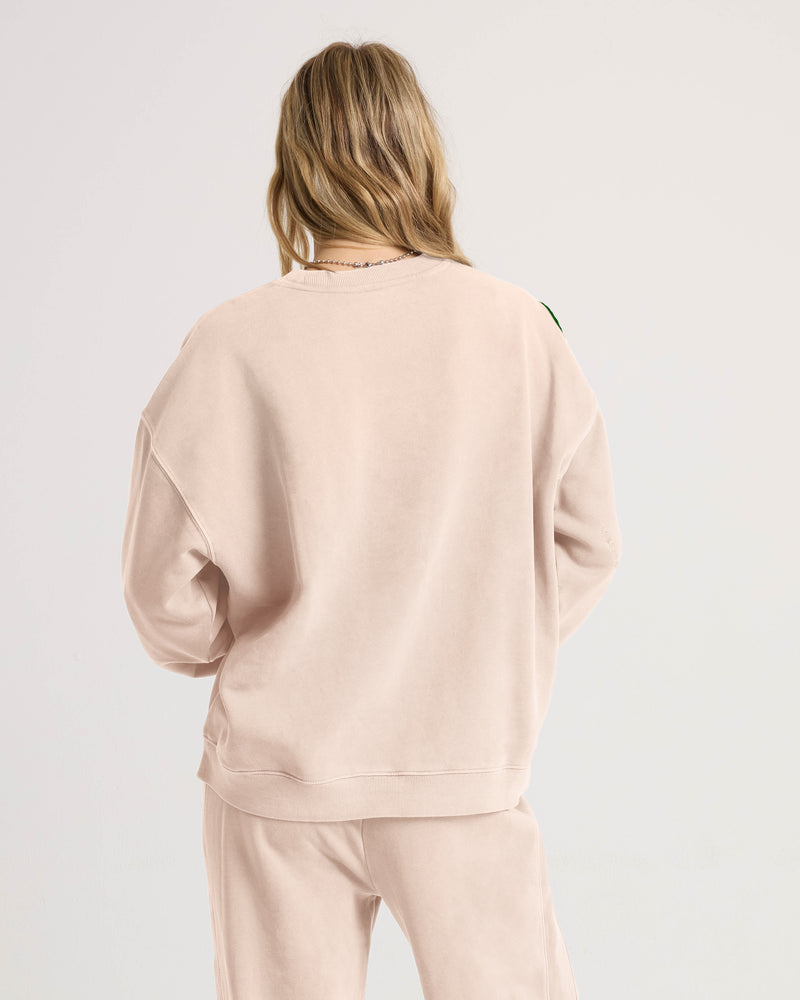 A woman with long, wavy blonde hair is seen from behind wearing Volcom’s Women’s Stone Soft Crew in light beige and matching slub brushed back fleece pants, standing against a plain, light background for that signature Lived In Lounge comfort.