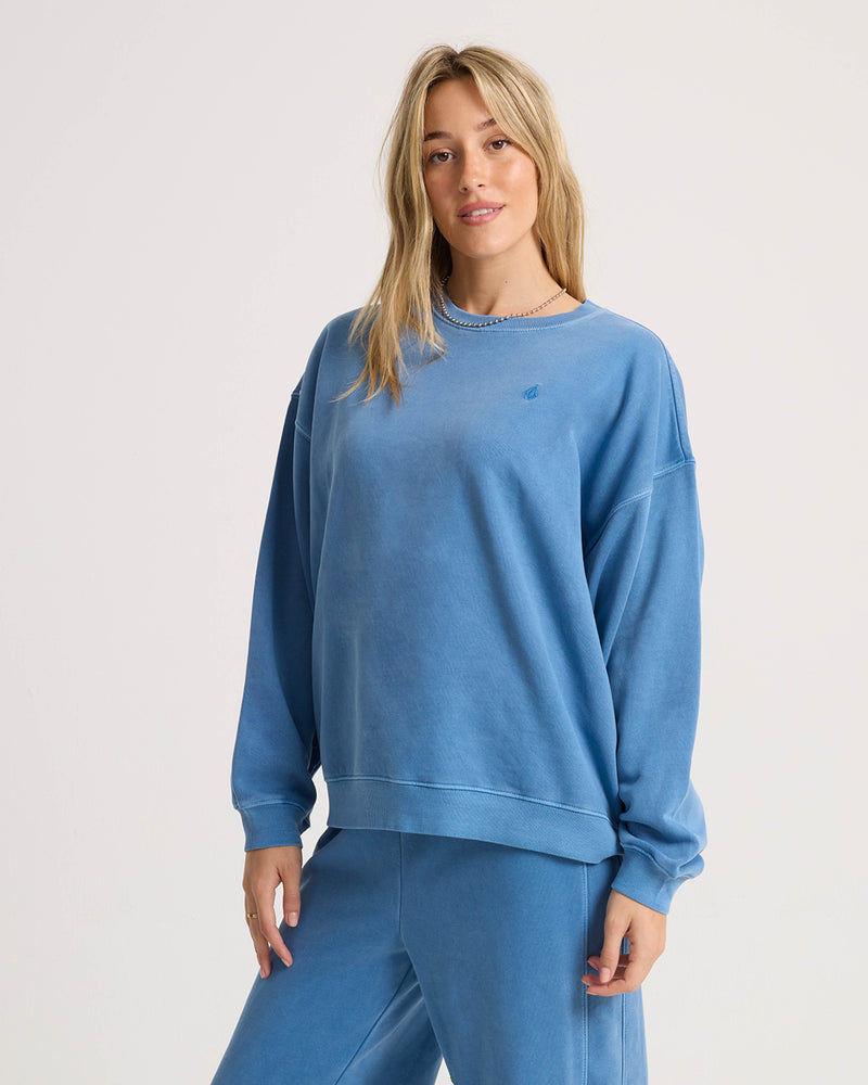 A woman wears a relaxed dropped-shoulder blue Volcom Women’s Stone Soft Crew sweatshirt with a small embroidered logo on the chest, paired with matching sweatpants.