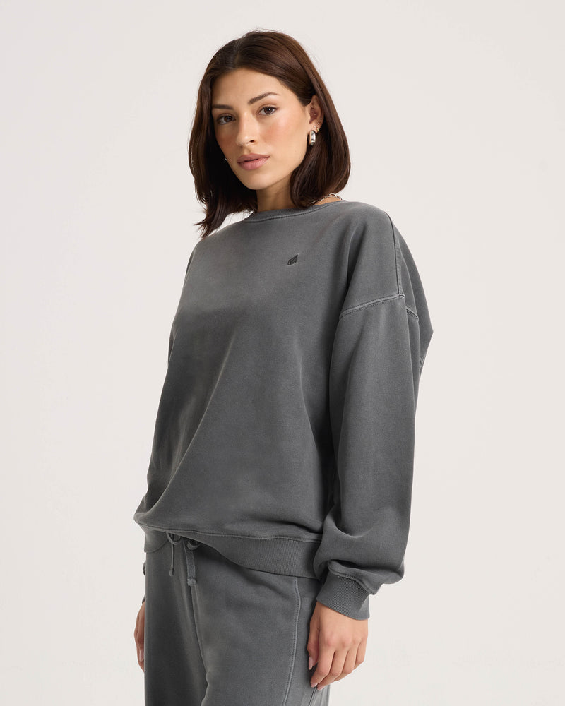 A woman wearing the Volcom Women’s Stone Soft Crew—a relaxed fit, long-sleeved gray sweatshirt crafted from soft fleece—along with matching sweatpants stands against a plain light background, looking at the camera with a neutral expression.