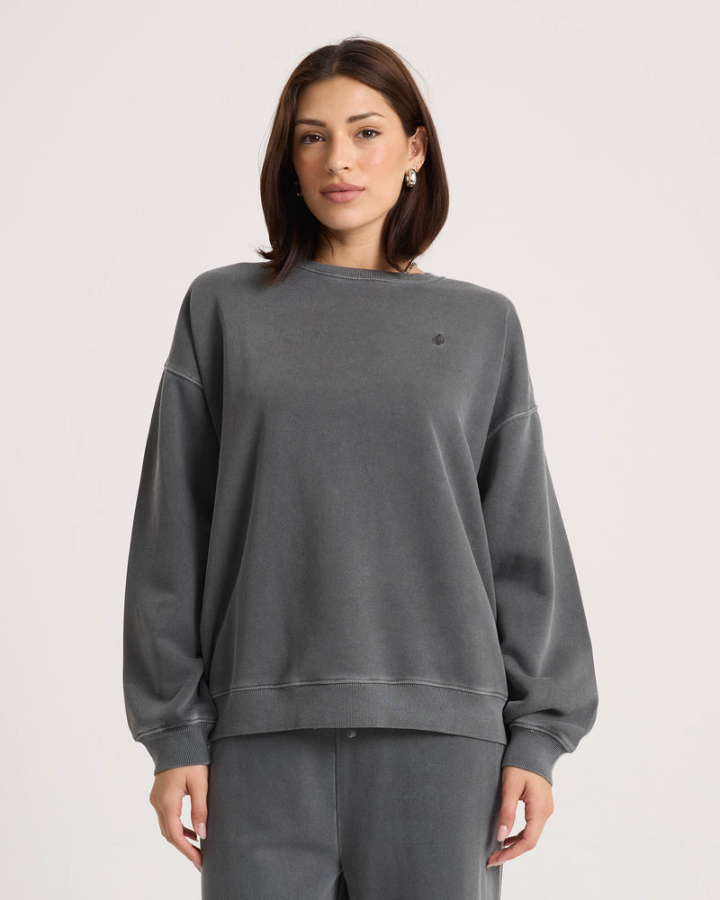 A woman with straight brown hair is wearing the Volcom Women’s Stone Soft Crew in a relaxed fit and dark gray color, along with matching soft fleece pants, as she stands against a plain light background.