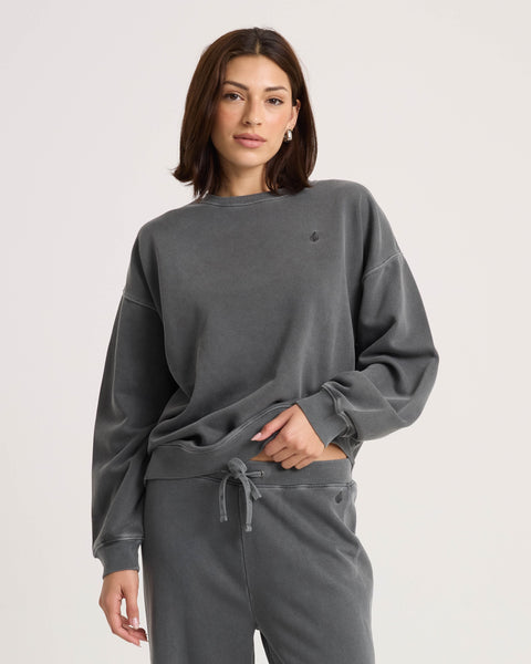 A person stands against a plain background, casually posing with one hand in their pocket and a relaxed expression, wearing the Volcom Women's Stone Soft Crew in dark gray along with matching pants. The soft fleece fabric of the sweatshirt enhances the comfortable and laid-back vibe of the outfit.