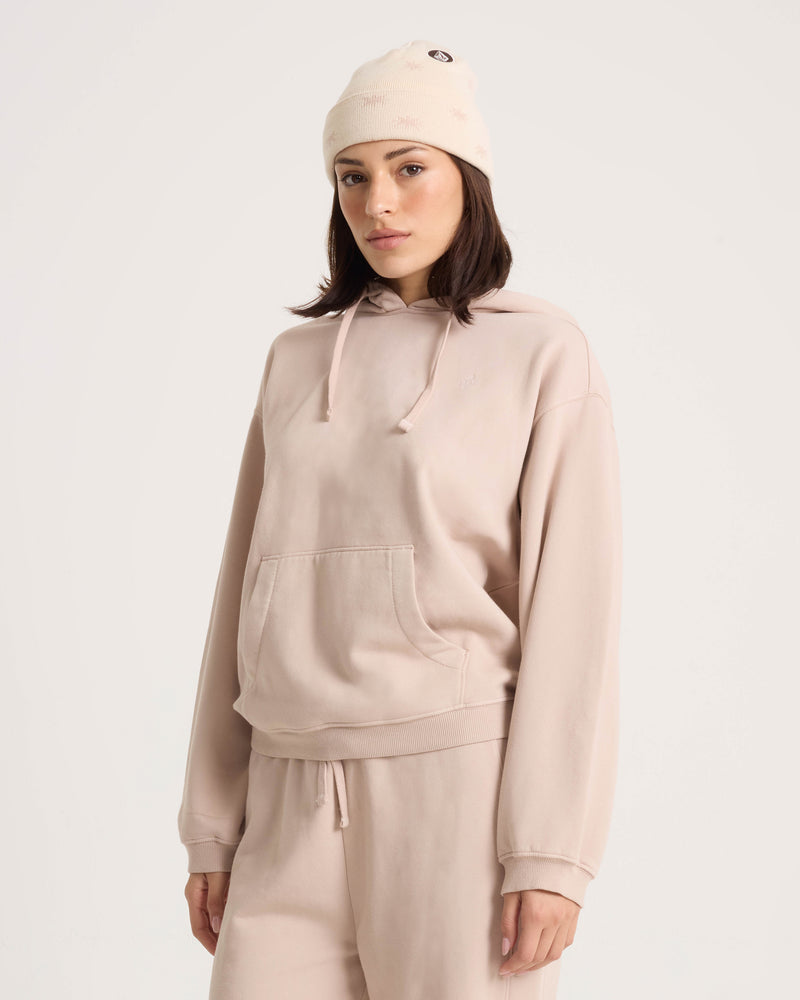 A woman wearing the Volcom Women’s Stone Soft Hoodie in beige stands against a plain light background, looking at the camera with a neutral expression. She pairs the cotton-blend fleece hoodie with matching sweatpants and a knit beanie.