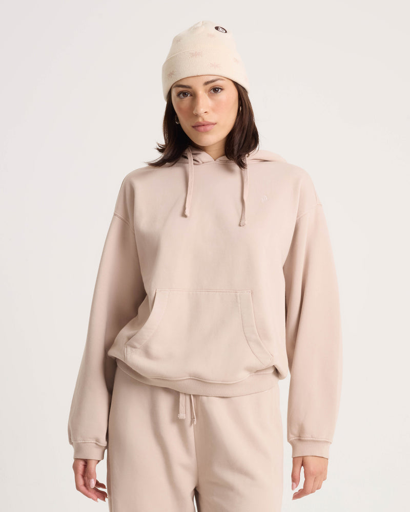 A woman wearing the Volcom Women’s Stone Soft Hoodie in light beige cotton-blend fleece, along with matching sweatpants and a cream knit beanie, stands against a plain white background, looking at the camera with a neutral expression.