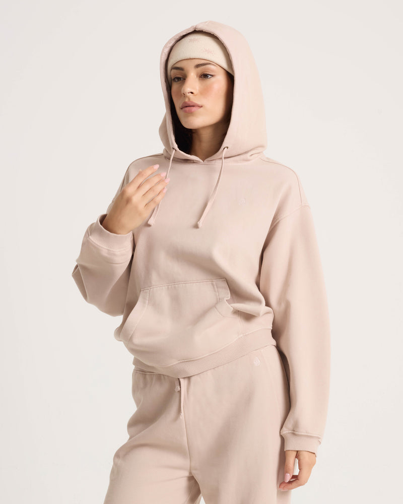 Wearing the Volcom Women’s Stone Soft Hoodie and matching sweatpants, crafted from cotton-blend fleece, a person stands against a plain background with one hand near their face and a neutral expression.