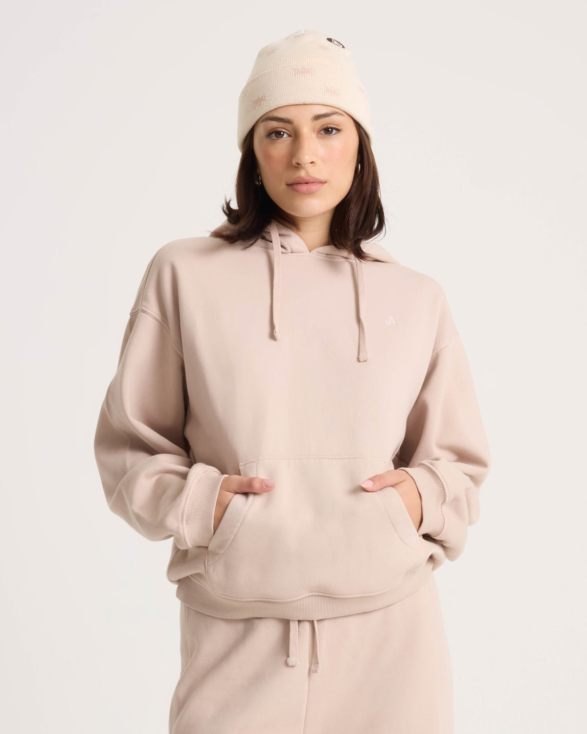 A woman stands with her hands in the pocket of the Volcom Women’s Stone Soft Hoodie in light beige, paired with a matching beanie, looking at the camera against a plain background.