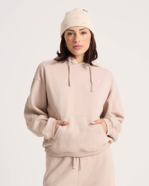 A woman stands with her hands in the pocket of the Volcom Women’s Stone Soft Hoodie in light beige, paired with a matching beanie, looking at the camera against a plain background.