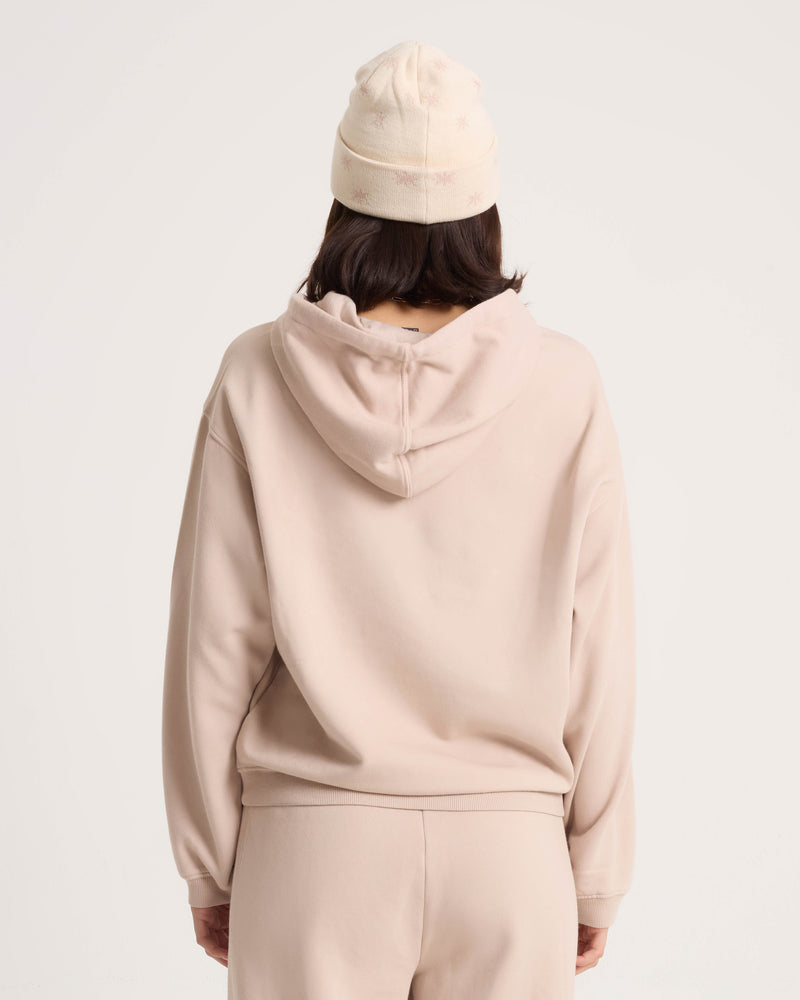A person stands facing away from the camera against a plain light background, wearing the Volcom Women’s Stone Soft Hoodie and matching pants, complemented by a cream-colored beanie. The hoodie’s cotton-blend fleece fabric introduces subtle texture to the understated look.