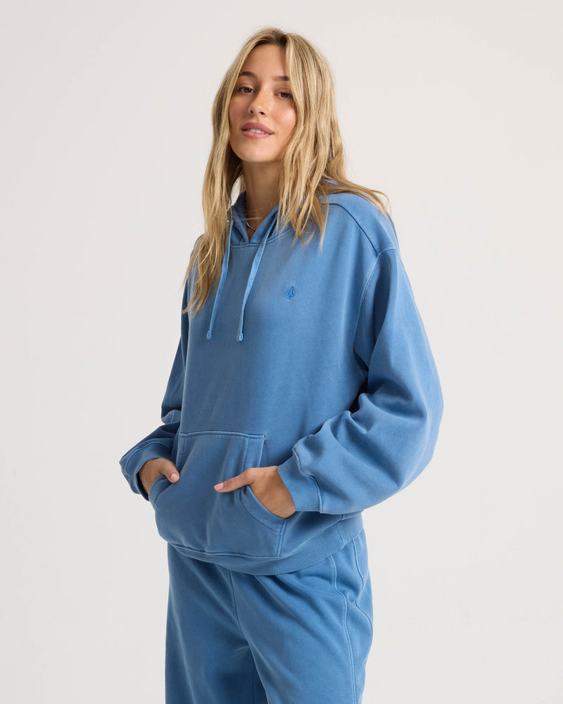 A woman wears a relaxed-fit, blue cotton-blend Volcom Women’s Stone Soft Hoodie, featuring a front kangaroo pocket and a small matching logo on the chest.