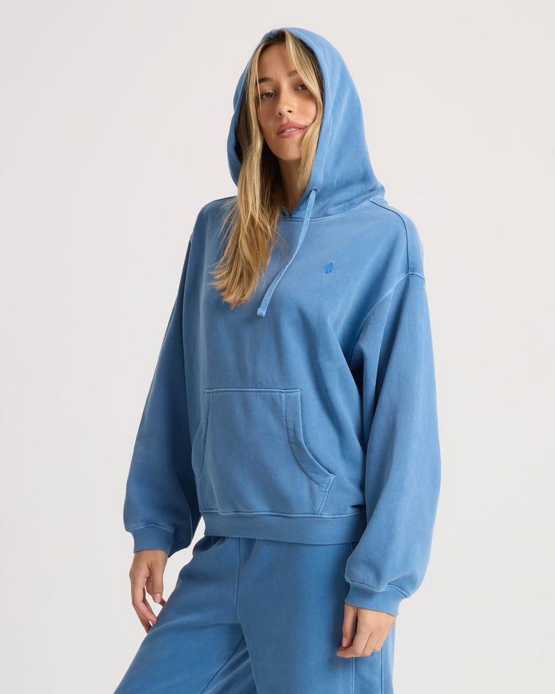 A woman wears a relaxed fit blue Volcom Women’s Stone Soft Hoodie with the hood up and a front kangaroo pocket.