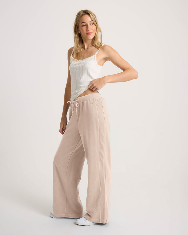 A woman wears a white ribbed tank top and Volcom Women’s Stone Soft Pant, which are light pink, loose-fit fleece sweatpants.