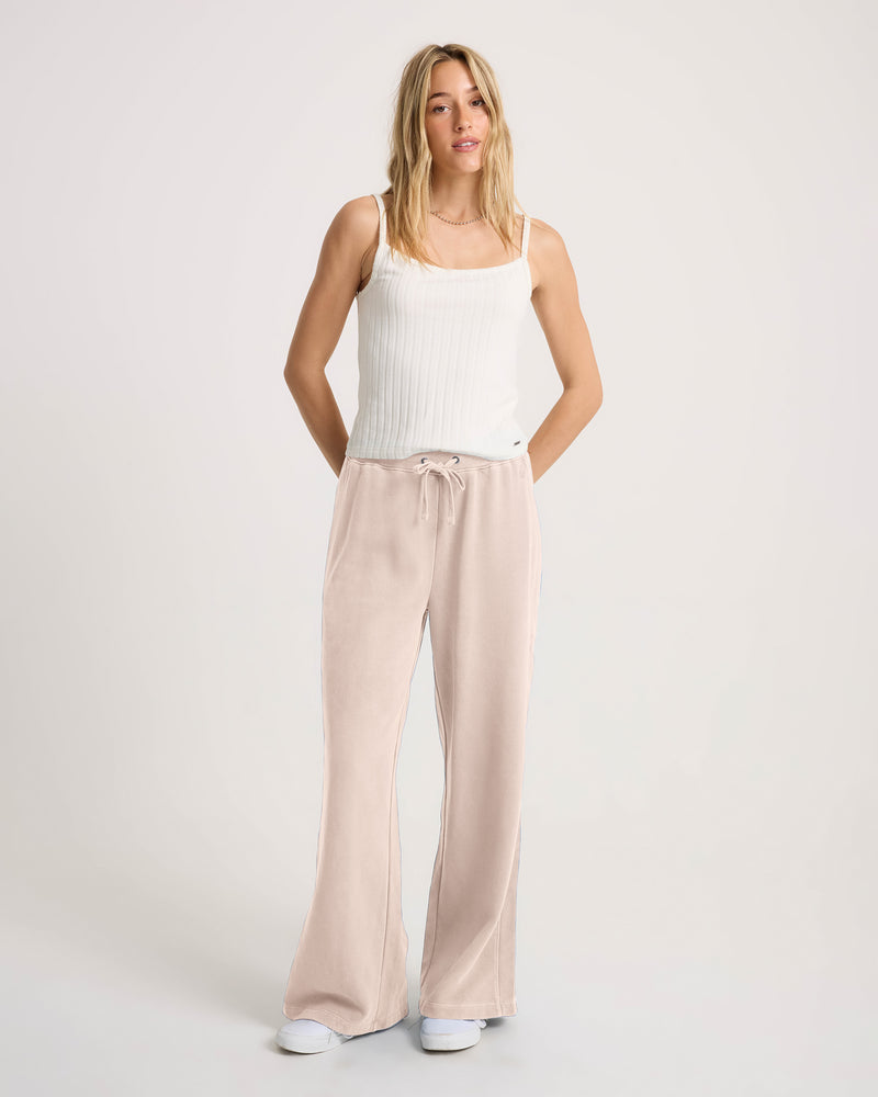 A woman wearing a white ribbed tank top and Volcom Women’s Stone Soft Pants in pale pink with a drawstring waist.