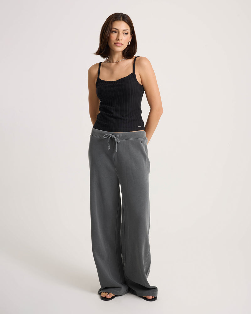 A woman with straight dark hair and a neutral expression stands against a plain background wearing a black tank top, Volcom Women’s Stone Soft Pants—crafted from a cotton-blend knit in a relaxed fit with a drawstring waist—and black slide sandals.