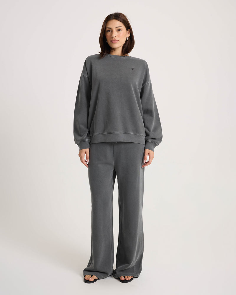 A woman stands facing forward, wearing a loose-fitting charcoal gray cotton-blend knit sweatshirt and Volcom Women’s Stone Soft Pants in a relaxed wide-leg fit, paired with black sandals against a plain white background.