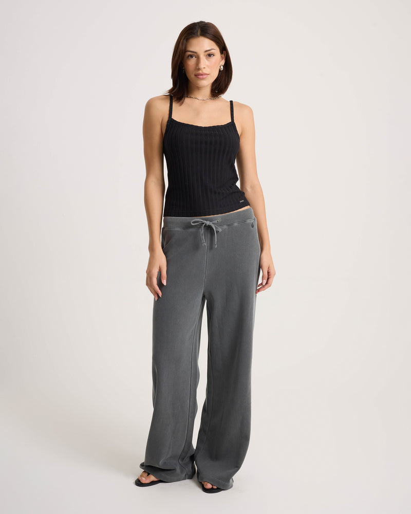 A woman stands against a plain background wearing a black sleeveless top, Volcom Women’s Stone Soft Pants in relaxed-fit gray with a drawstring, and black sandals.