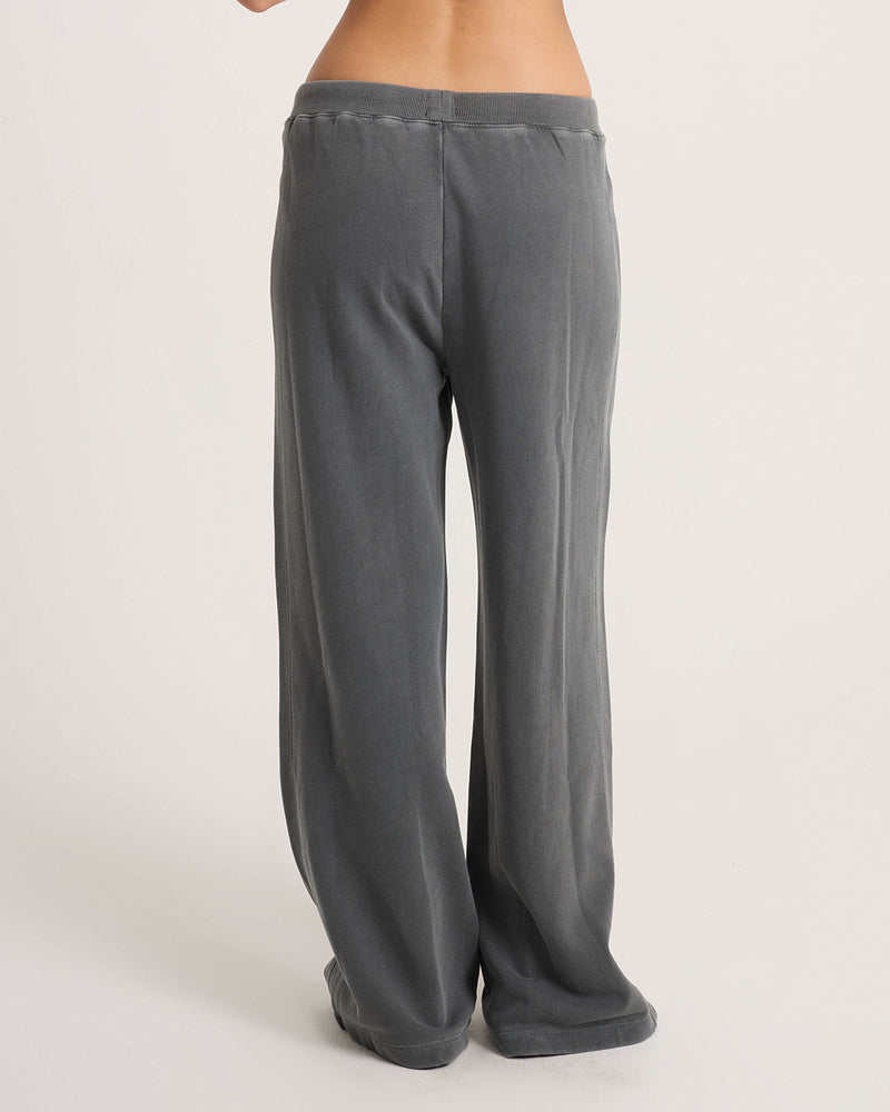 A person is shown from the back, wearing Volcom Women’s Stone Soft Pants—loose-fitting, high-waisted gray cotton-blend knit sweatpants in a relaxed fit. Only their lower back and legs are visible as they stand against a plain off-white background.