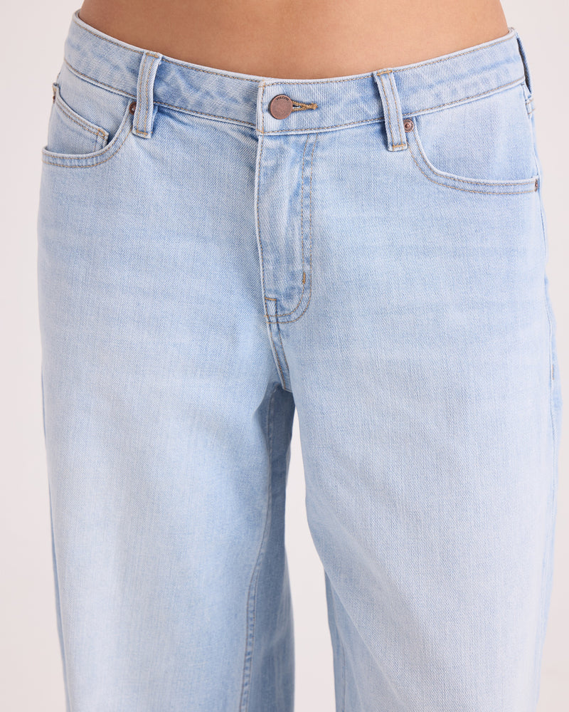 A close-up of a person wearing Volcom Women’s Stoned BF Lowrise Pants in light blue denim, made from stretch cotton, highlighting the waistband, button, zipper, and upper thigh area against a plain background.