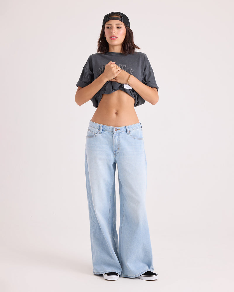 A person wearing a backwards black cap, a gray crop top, and Volcom Women’s Stoned BF Lowrise Pants stands against a plain light background, hands clasped at chest.