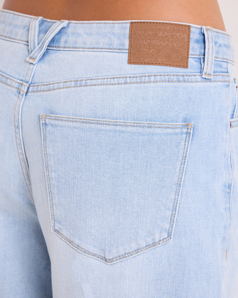 Close-up of the back pocket and waistband of the Volcom Women’s Stoned BF Lowrise Pants in light blue denim, highlighting a brown leather patch with stitched detailing near the belt loop. Made from stretch cotton, these lowrise wide-leg jeans offer classic 5-pocket styling for comfort and versatility.