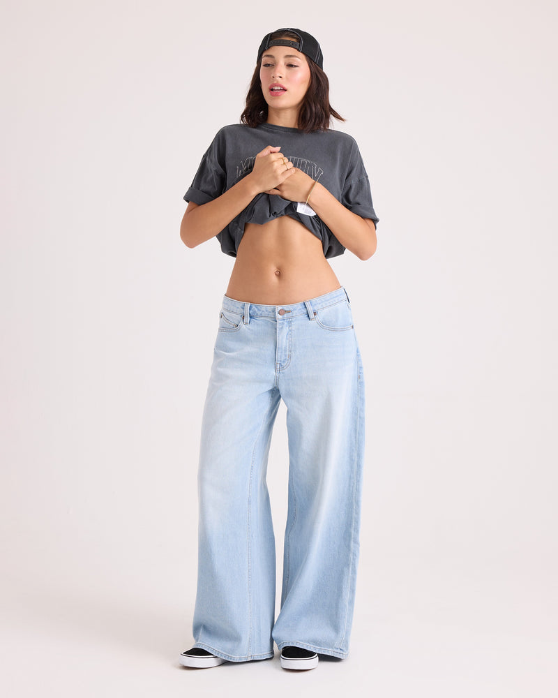 A woman stands against a plain background, wearing a gray stretch cotton shirt and Volcom Women’s Stoned BF Lowrise Pants—light blue, wide-leg jeans with 5-pocket styling—paired with sneakers. She lifts her shirt to reveal her midriff, wears a backwards cap, and gazes slightly upward.