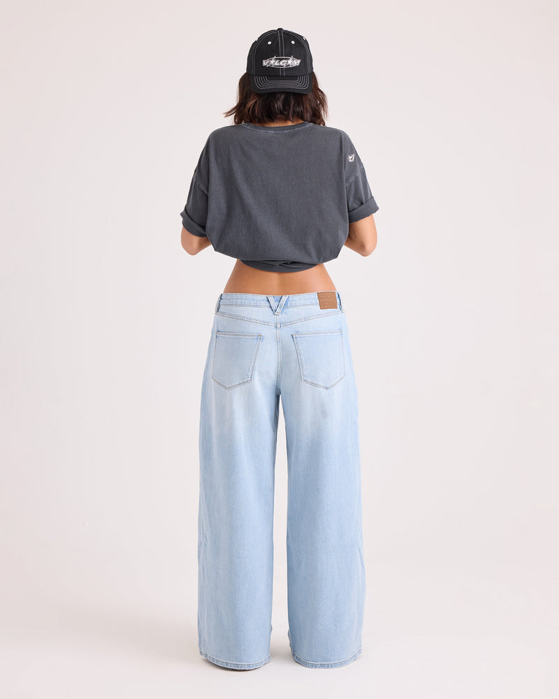 A person stands facing away, wearing a black cap, a dark cropped t-shirt, and Volcom Women’s Stoned BF Lowrise Pants in light blue—a pair of wide-leg jeans crafted from stretch cotton with visible back pockets and a brown patch on the waistband. The background is plain and light-colored.