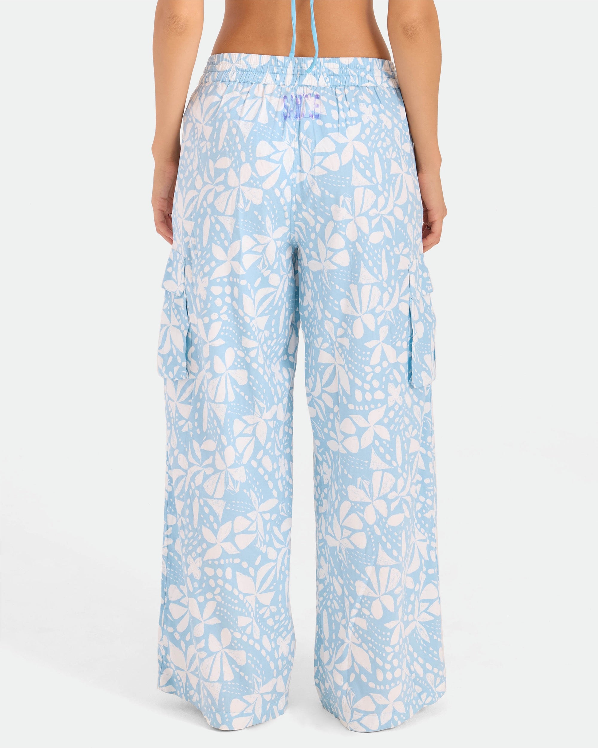 A person is seen from the back wearing Volcom Women's Wide Leg Pull On Pants in high-waisted light blue with a white floral and leaf print, soft cotton-viscose blend, side cargo pockets, elasticated waistband, and relaxed fit.